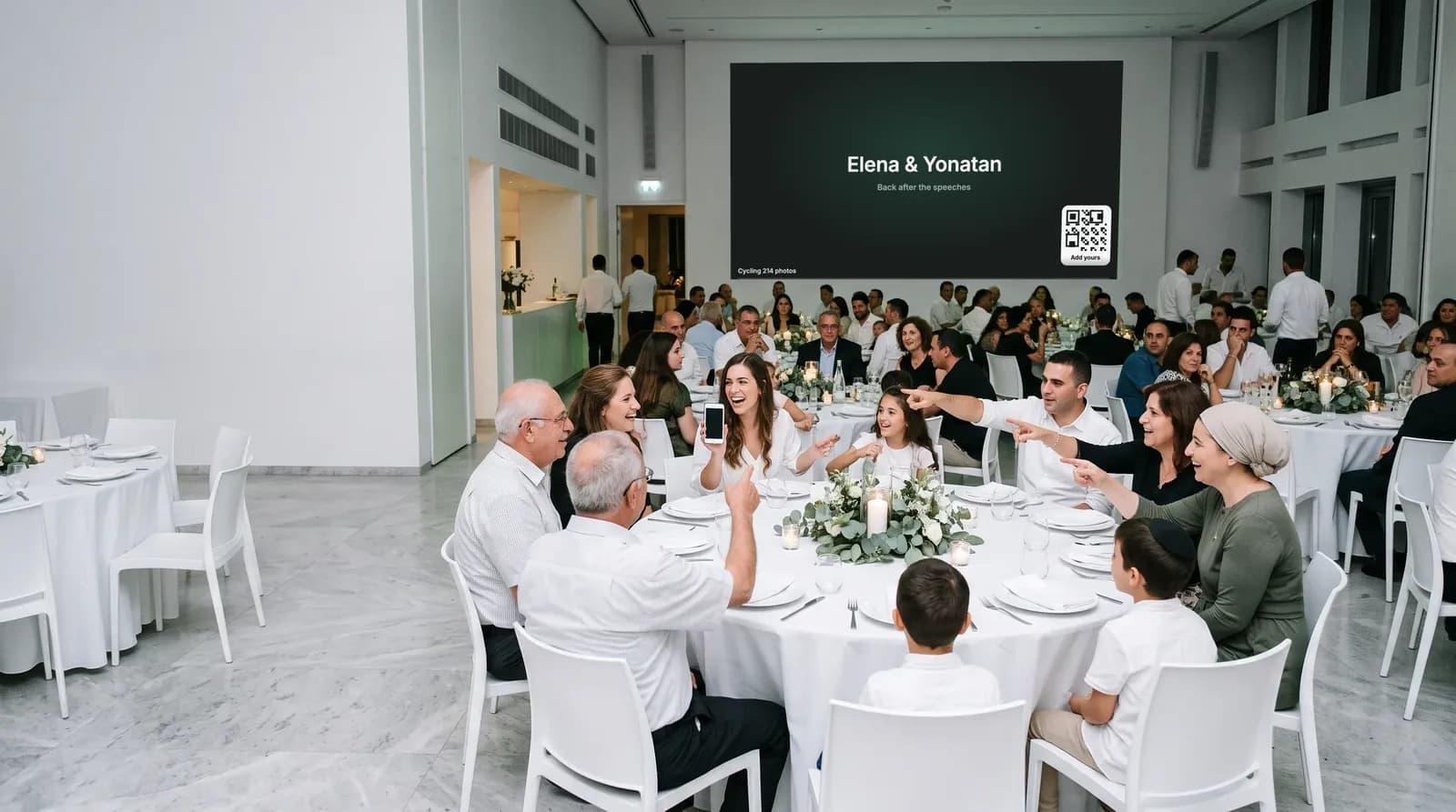 A white hall during dinner, guests at round tables turning towards a large screen on the wall that shows the couple’s names and a QR code while the photos pause for the speeches