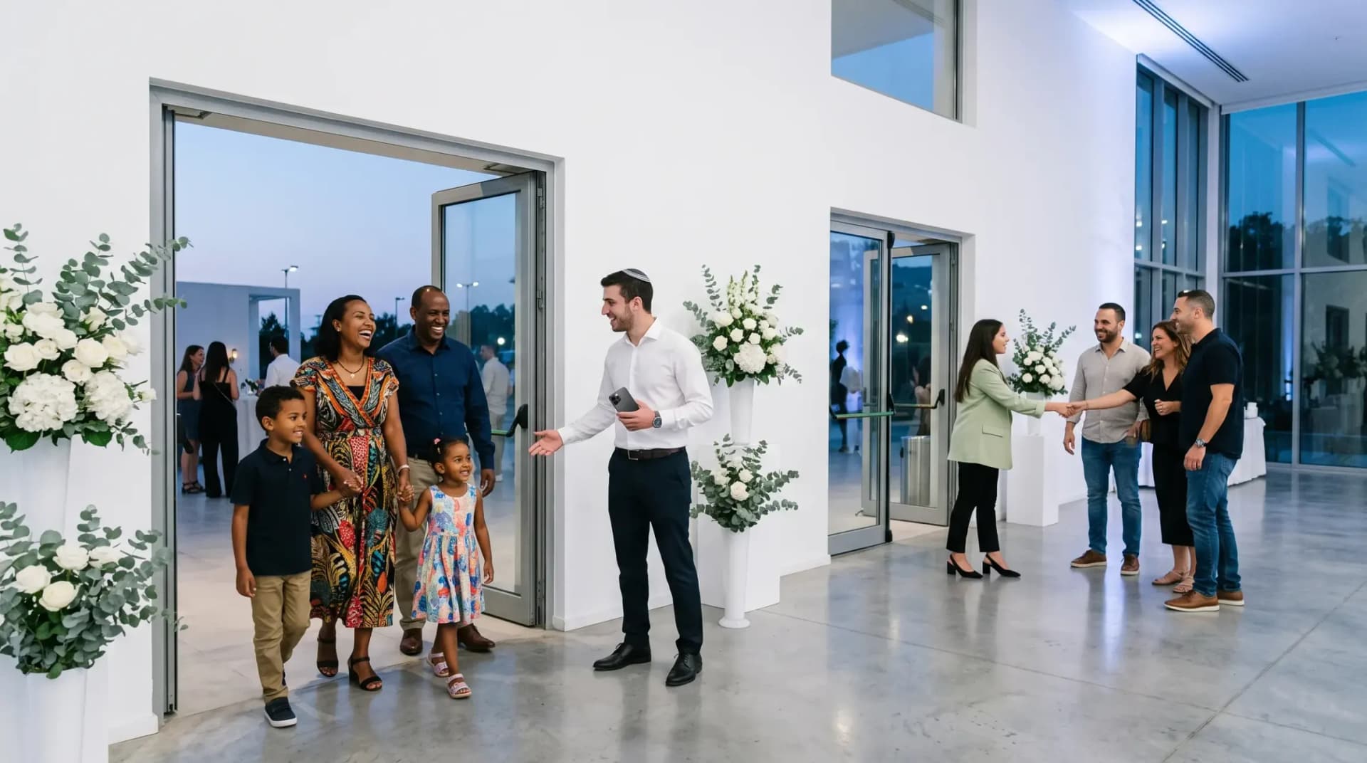 A family with two children is welcomed at one entrance of a bright event hall, while a host greets friends at a second entrance.