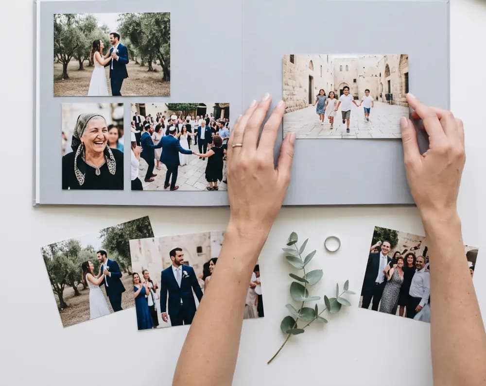 ידיים מסדרות תמונות חתונה מודפסות על כפולה אפורה בהירה של אלבום, ולידן הדפסות וענף אקליפטוס על שולחן לבן.
