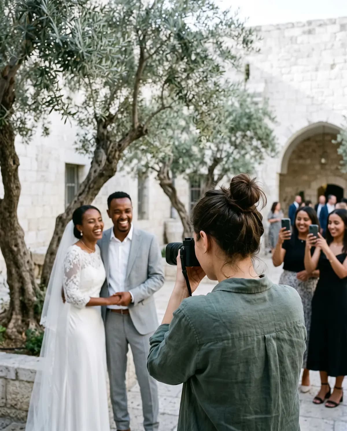 צלמת בחולצת פשתן מצלמת חתן וכלה צוחקים מתחת לעצי זית בחצר אבן, ושני אורחים מצלמים אותם בטלפון.
