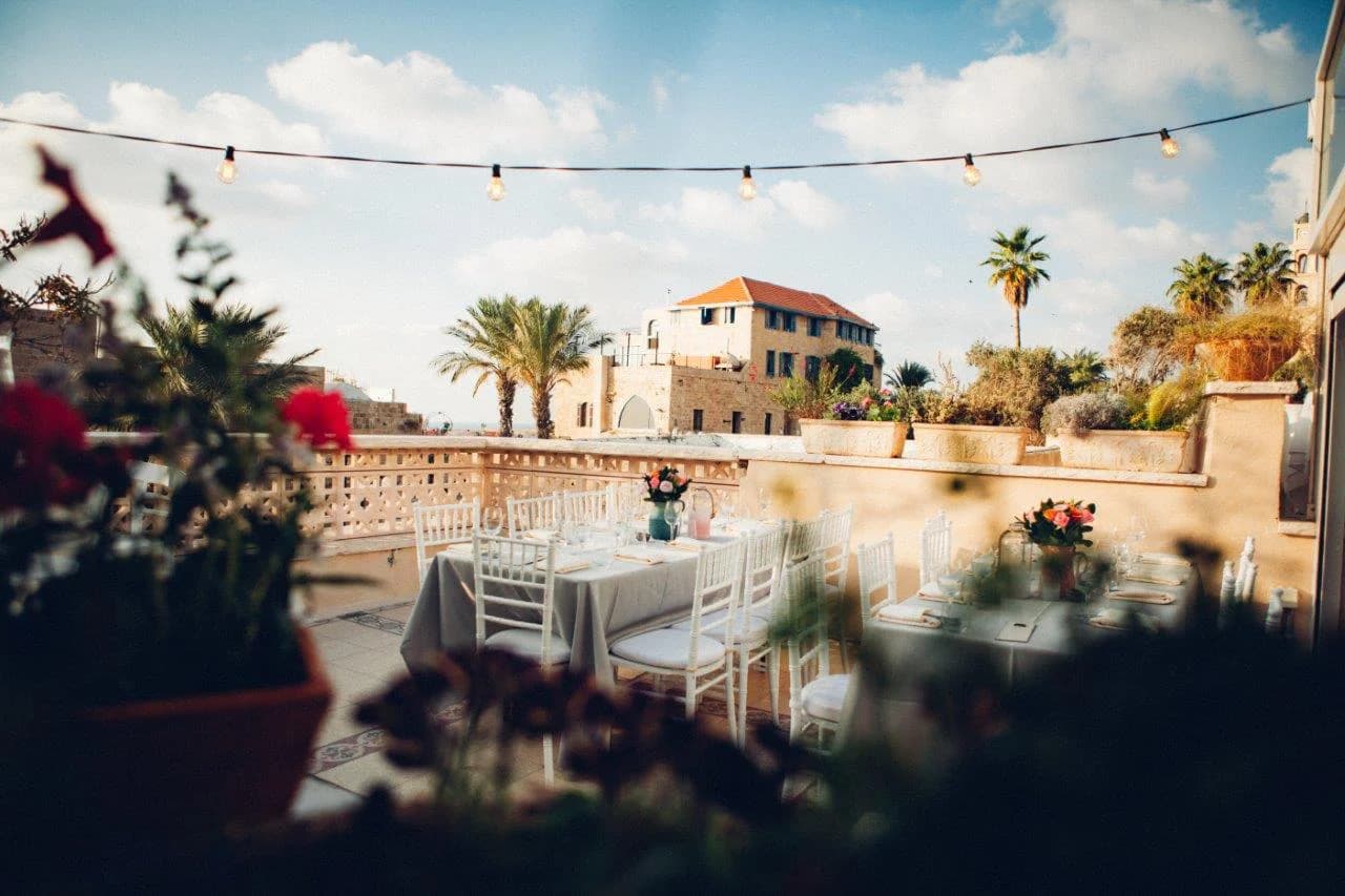 Terrace at Otzarin with white wooden chairs, string lights, palm trees and a glimpse of the sea