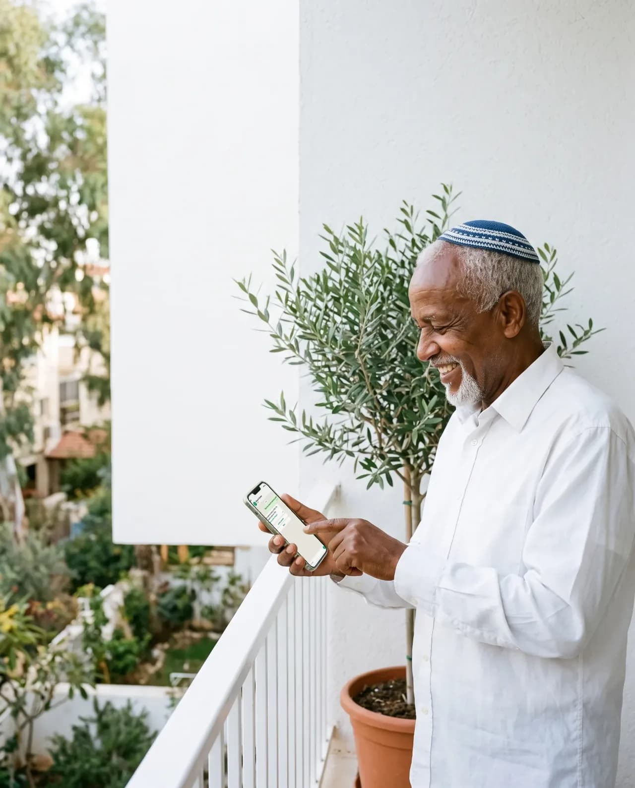 An older man with a kippah on a white balcony smiling at his phone