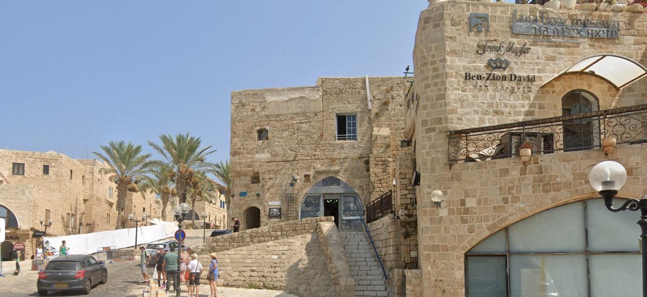 Street view of the stone-arched entrance to Otzarin in Old Jaffa, with steps leading up and palm trees nearby