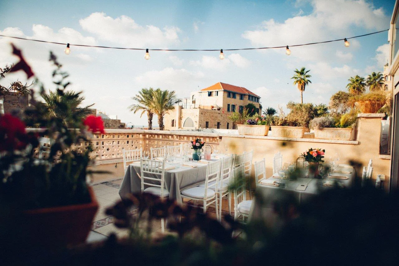 Terrace at Otzarin with white wooden chairs, string lights, palm trees and a glimpse of the sea